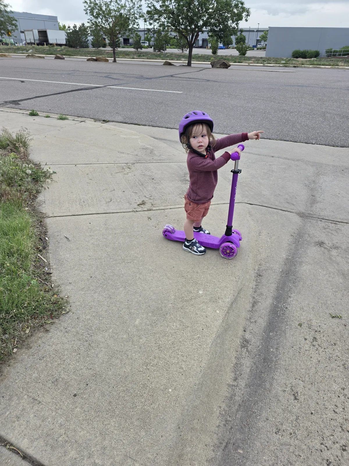 Xander in a purple helmet on his purple scooter at a curb, pointing down the street toward the park.