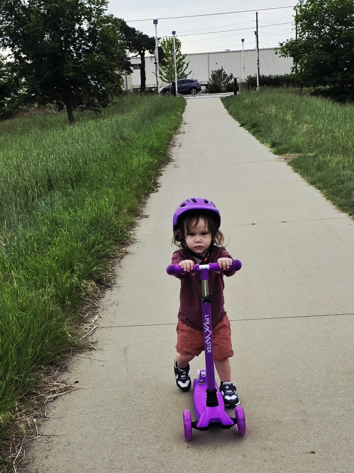 Xander pushing the purple scooter up a long sidewalk, head down, industrial buildings behind him.