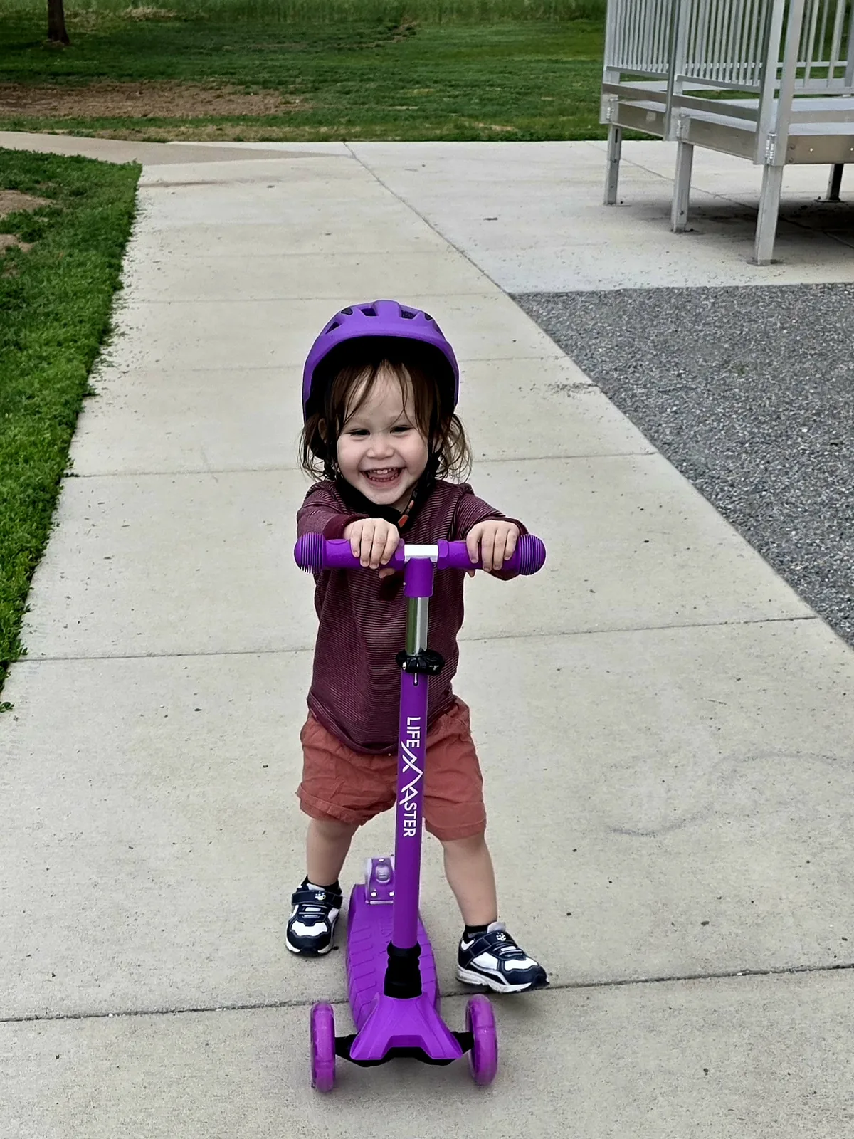 Xander grinning at the camera with both hands on his purple scooter, at the school by the park.